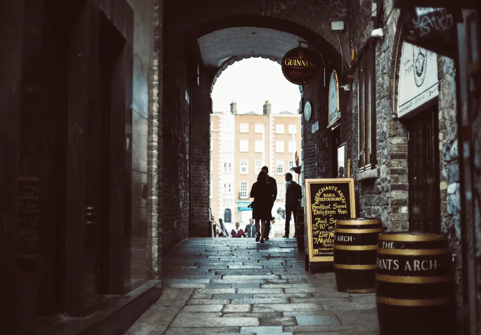 A narrow, stone-paved alleyway with arched ceilings leads to a bright opening revealing buildings, with two people walking away from the viewer, and a 'Merchants Arch' sign on the right.