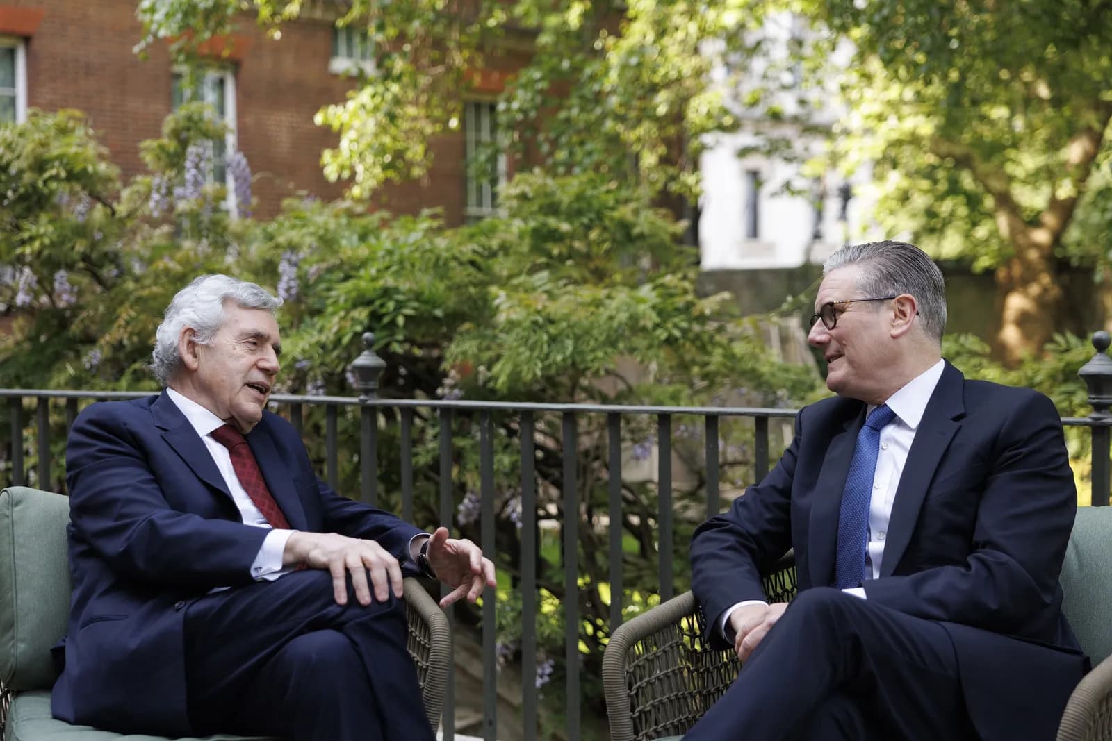 Two men in suits are seated outdoors, conversing, with greenery and a building in the background.